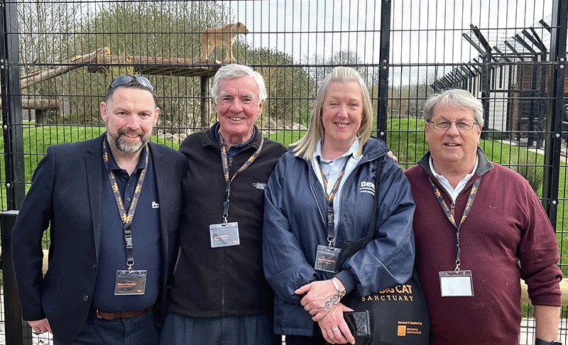 Steve Crawley, John Burton, Sarah Hashmi and Terry Capps pose in front of a lion enclosure, with the lion standing proudly on a raise platform in the background.