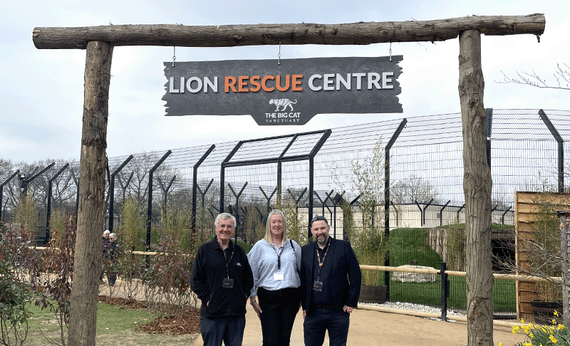 John Burton, Sarah Hashmi and Steve Crawley (l-r) standing in front of a large wooden sign featuring the words 'Lion Rescue Centre' in white and orange writing.