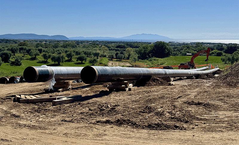 Two long, large diameter pipes resting on the floor against the backdrop of green fields and blue sky.