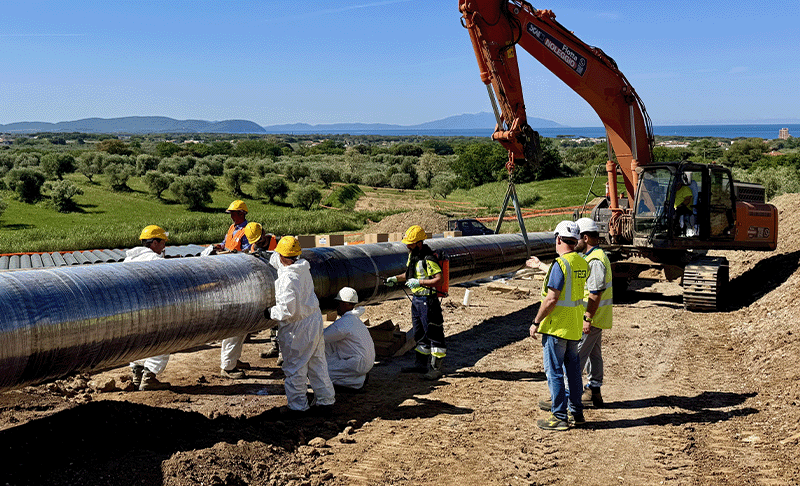 Machinery holding up a long piece of pipe, with many people applying tapes around the circumference of the pipe, set against a backdrop of green fields and blue sky.